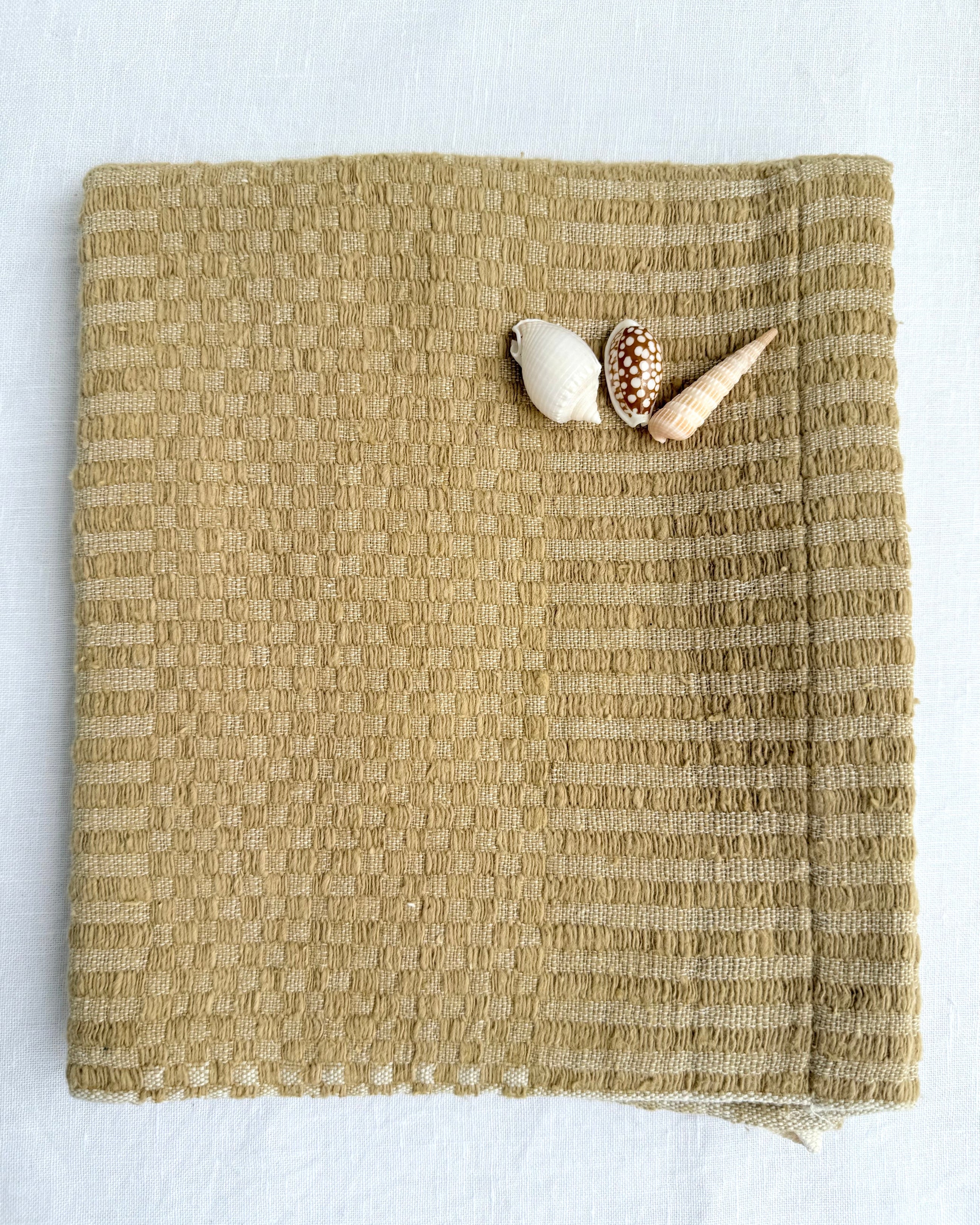 A folded Bronze-coloured woven hand towel displayed on a white background with three small seashells placed on the upper right corner.
