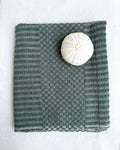 A folded teal-colored hand towel with a subtle checker pattern, displayed on a white fabric background with a white sea urchin shell used as a size reference.