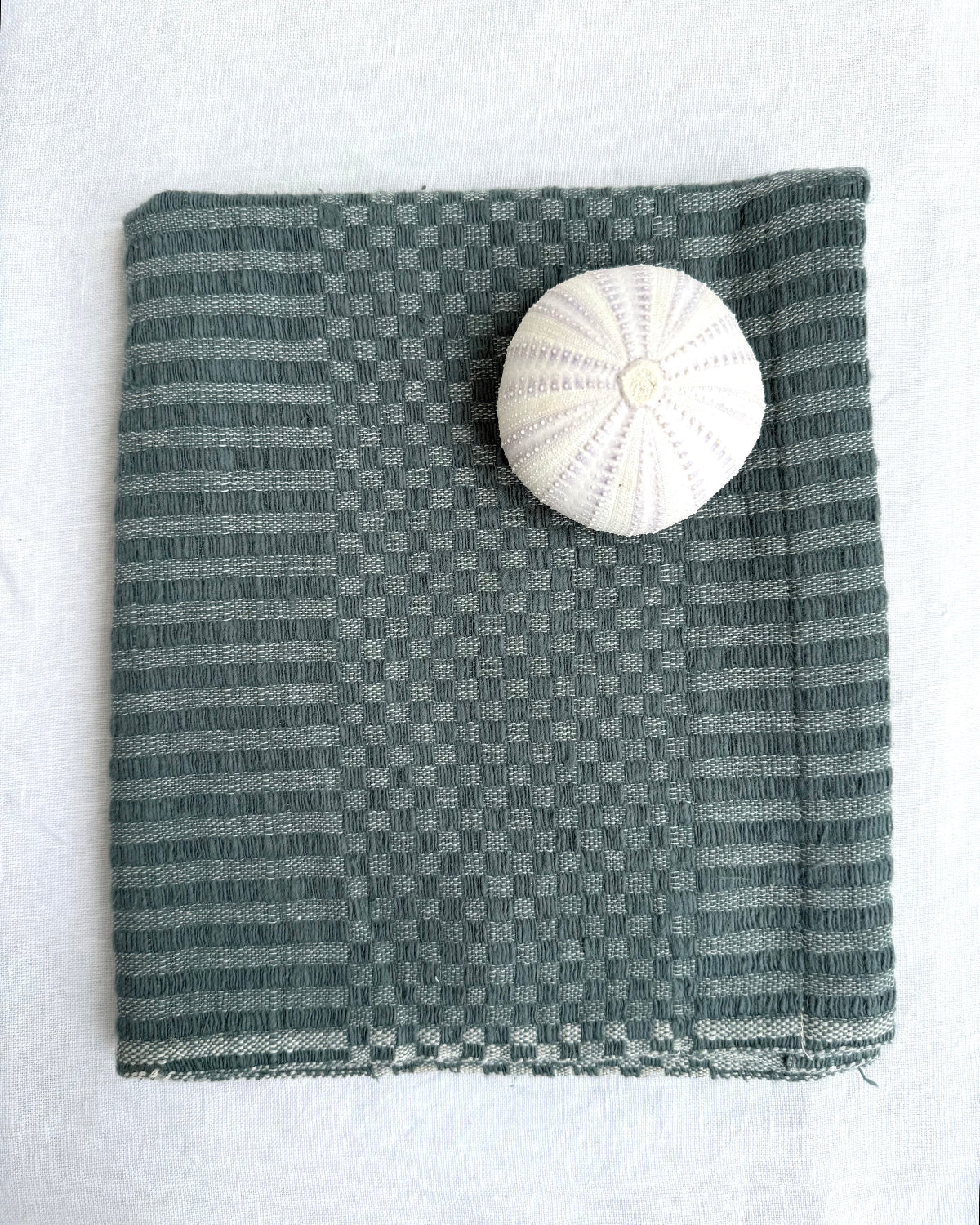 A folded teal-colored hand towel with a subtle checker pattern, displayed on a white fabric background with a white sea urchin shell used as a size reference.