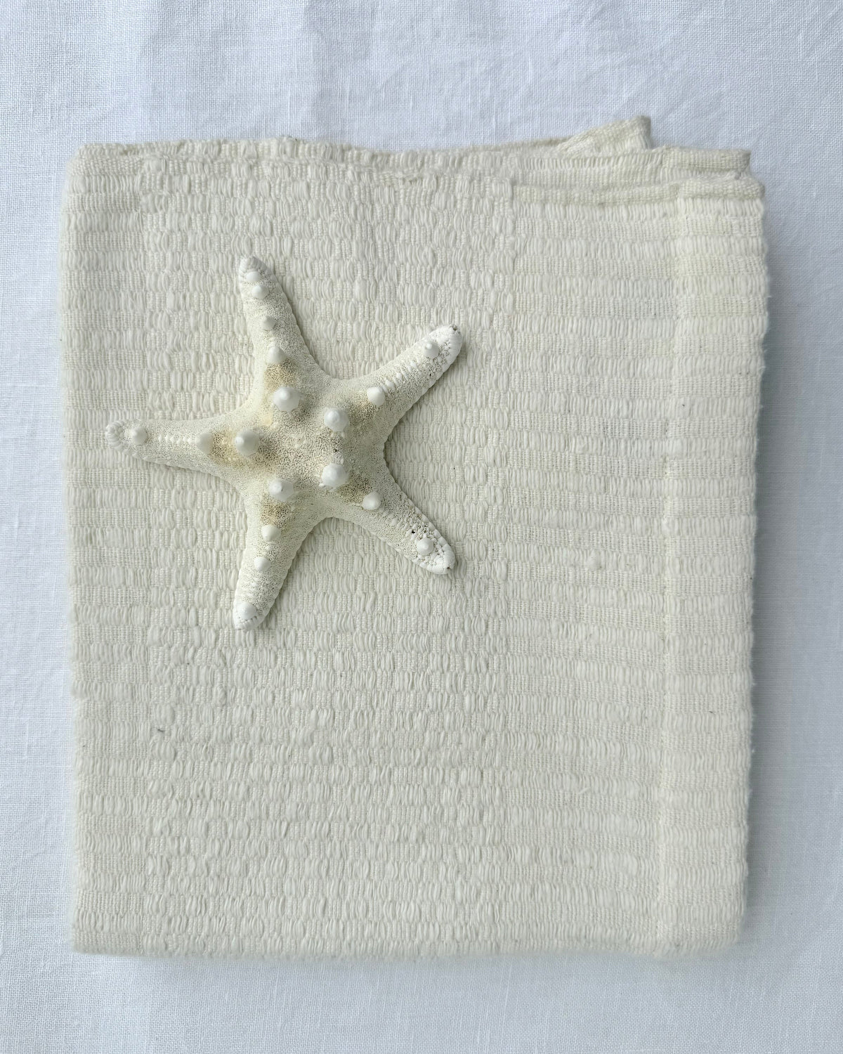 A folded cream-colored hand towel, featuring a small white starfish decoration on the front, set against a matching white fabric background.
