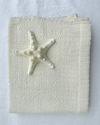 A folded cream-colored hand towel, featuring a small white starfish decoration on the front, set against a matching white fabric background.