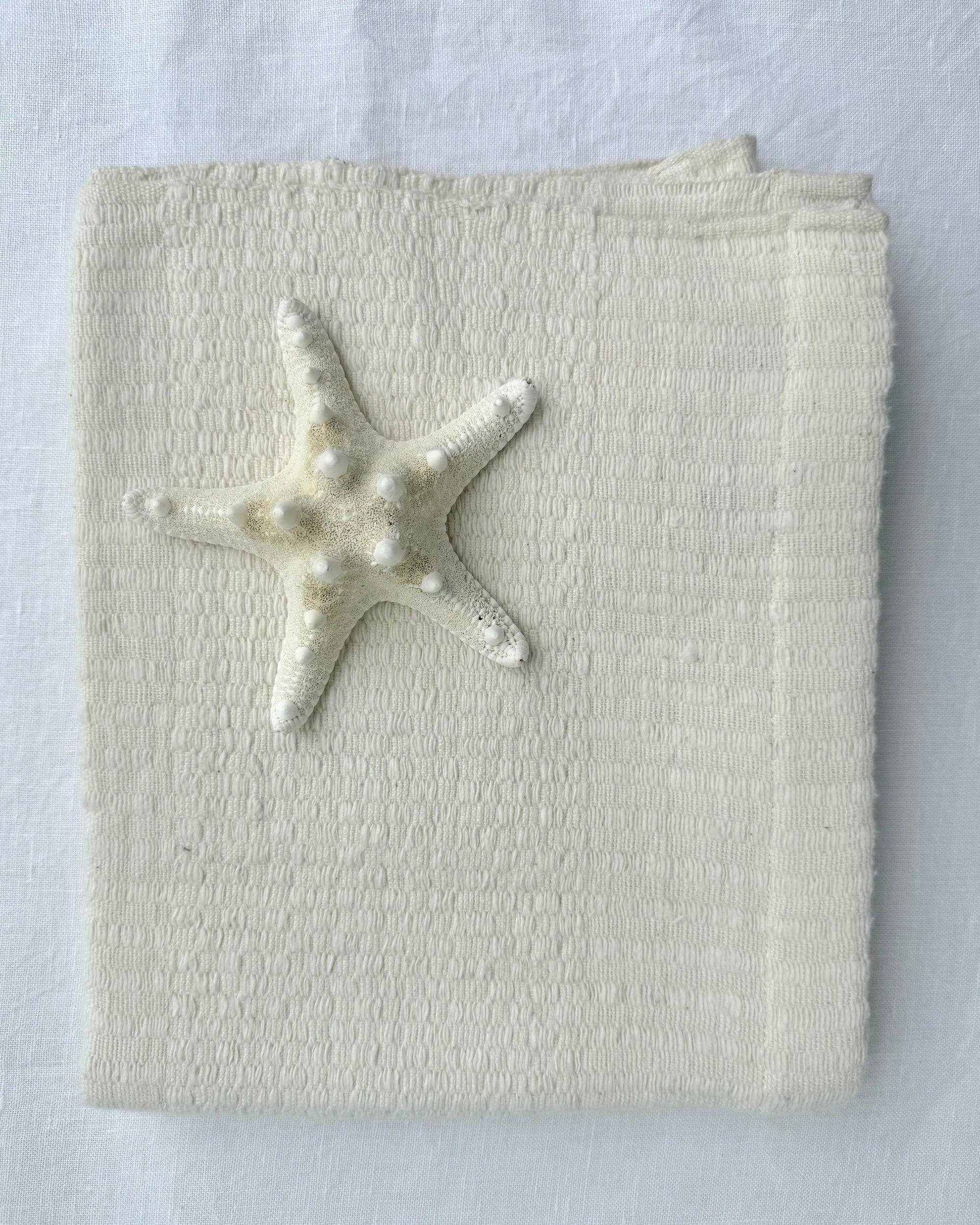 A folded cream-colored hand towel, featuring a small white starfish decoration on the front, set against a matching white fabric background.