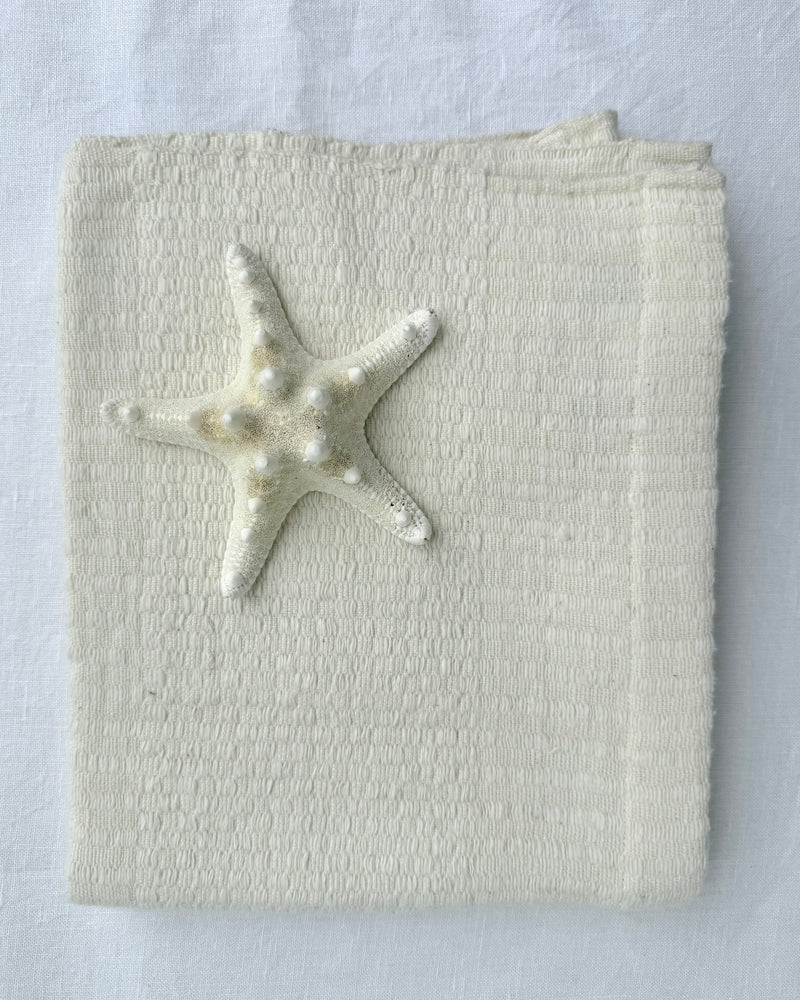 A folded cream-colored hand towel, featuring a small white starfish decoration on the front, set against a matching white fabric background.