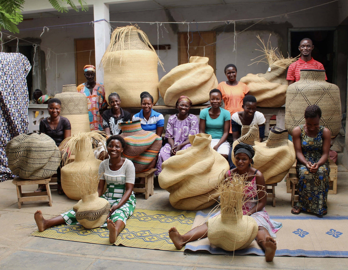 A group of women posing with large, hand-woven natural fiber baskets and gourds in an outdoor workshop setting.