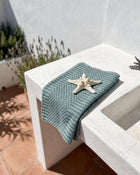 A folded sage green textured cotton towel with a white starfish on top, styled on a white concrete outdoor sink against a white wall.