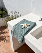 A folded sage green textured cotton towel with a white starfish on top, styled on a white concrete outdoor sink against a white wall.