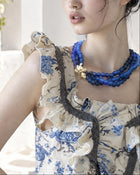 A woman with dark hair in a messy bun wearing a cream and blue floral ruffled top and a blue beaded necklace, posing outdoors in front of a white archway with green plants.