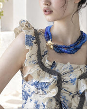 A woman with dark hair in a messy bun wearing a cream and blue floral ruffled top and a blue beaded necklace, posing outdoors in front of a white archway with green plants.
