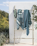 Two teal beach towels draped over a rustic wooden ladder outdoors against a white wall in an outdoor bathroom.