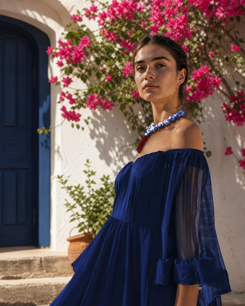 A woman wearing a royal blue off-the-shoulder dress and a matching blue beaded necklace, standing outdoors in front of a white wall with a blue door and bright pink bougainvillea flowers.