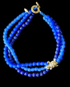 A beaded necklace made of multiple strands of round blue beads in varying shades, with a gold toggle clasp and a small gold decorative accent, shown on a solid black background.