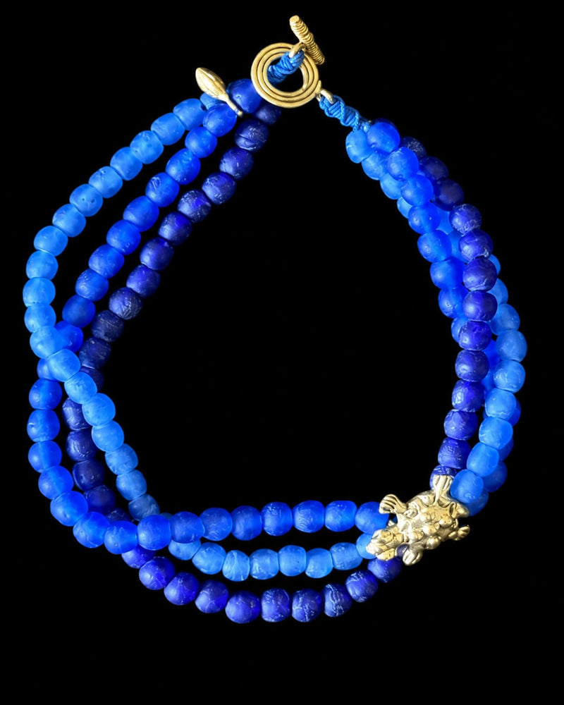A beaded necklace made of multiple strands of round blue beads in varying shades, with a gold toggle clasp and a small gold decorative accent, shown on a solid black background.