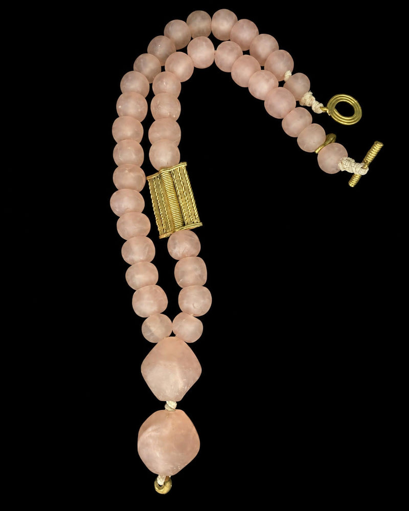 A rose quartz beaded necklace with a textured gold rectangular centerpiece and a toggle clasp, displayed on a solid black background.
