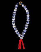 A beaded necklace or mala-style strand with white round beads decorated in blue floral patterns, a gold clasp at the top, and a red tassel hanging from the bottom center, shown against a solid black background.