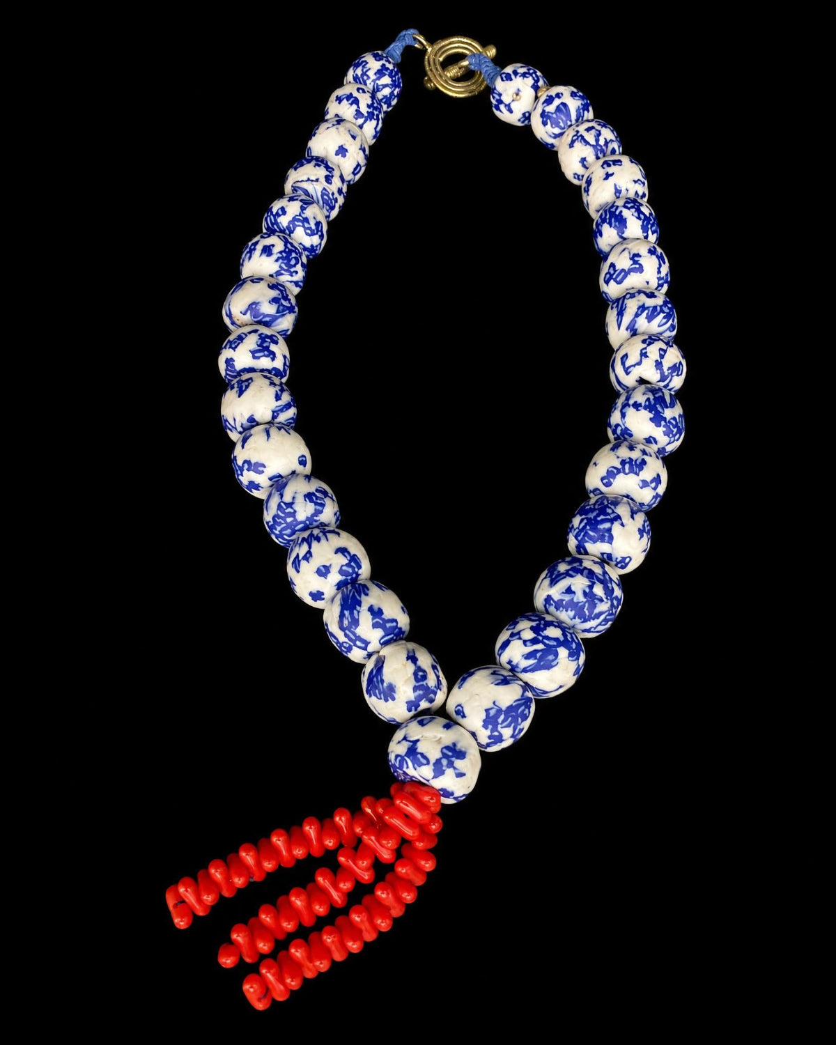 A beaded necklace featuring round blue and white patterned beads, a gold-tone clasp, and a dangling red beaded tassel, shown against a solid black background.