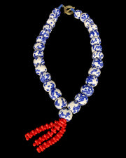 A beaded necklace featuring round blue and white patterned beads, a gold-tone clasp, and a dangling red beaded tassel, shown against a solid black background.