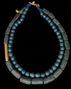 Ancient Egyptian style necklace featuring blue-green glass beads and dark gray stone cylinder beads, accented with gold-toned metal details and a blue cord clasp, shown against a solid black background.
