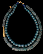 Ancient Egyptian style necklace featuring blue-green glass beads and dark gray stone cylinder beads, accented with gold-toned metal details and a blue cord clasp, shown against a solid black background.