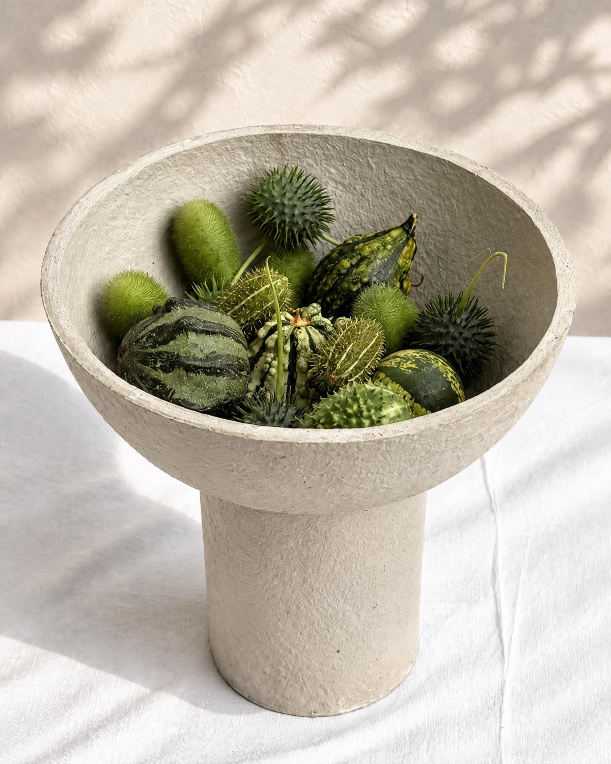 A textured paper pulp pedestal bowl filled with various green gourds and decorative botanical elements, set on a white fabric surface with soft leaf shadows in the background.