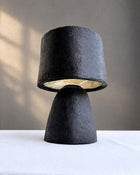 A sculptural table lamp with a textured black paper pulp base and a matching black fabric drum shade featuring a gold interior, set on a white tablecloth against a neutral gray wall.