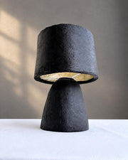 A sculptural table lamp with a textured black paper pulp base and a matching black fabric drum shade featuring a gold interior, set on a white tablecloth against a neutral gray wall.