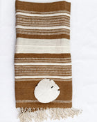 A folded brown and cream striped woven towel with fringe at the bottom, styled with a white sand dollar on a white background.