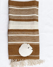 A folded brown and cream striped woven towel with fringe at the bottom, styled with a white sand dollar on a white background.