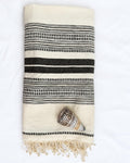 A folded cream-colored cotton towel with black woven stripes and fringe, styled with a small seashell on a white fabric background.