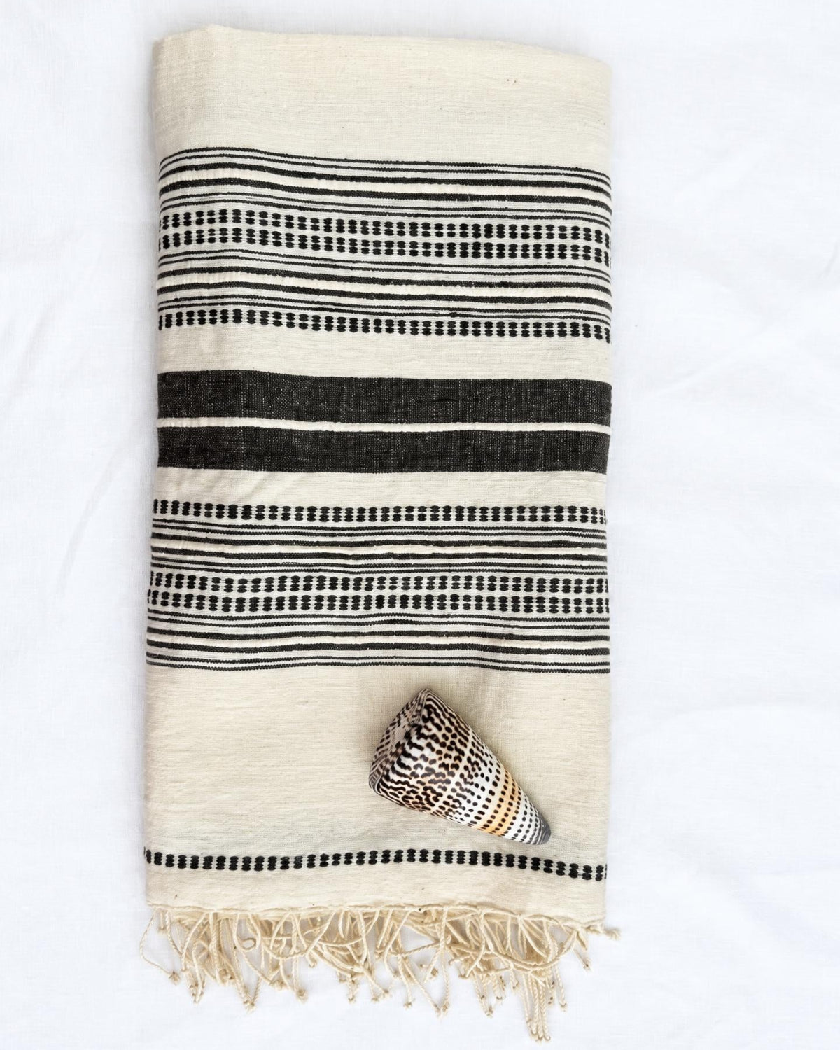 A folded cream-colored cotton towel with black woven stripes and fringe, styled with a small seashell on a white fabric background.