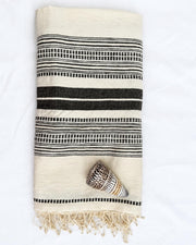 A folded cream-colored cotton towel with black woven stripes and fringe, styled with a small seashell on a white fabric background.