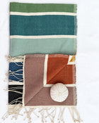 a woven towel laid flat on a white background. The larger textile at the top has horizontal stripes in olive green, teal, mint, and cream.