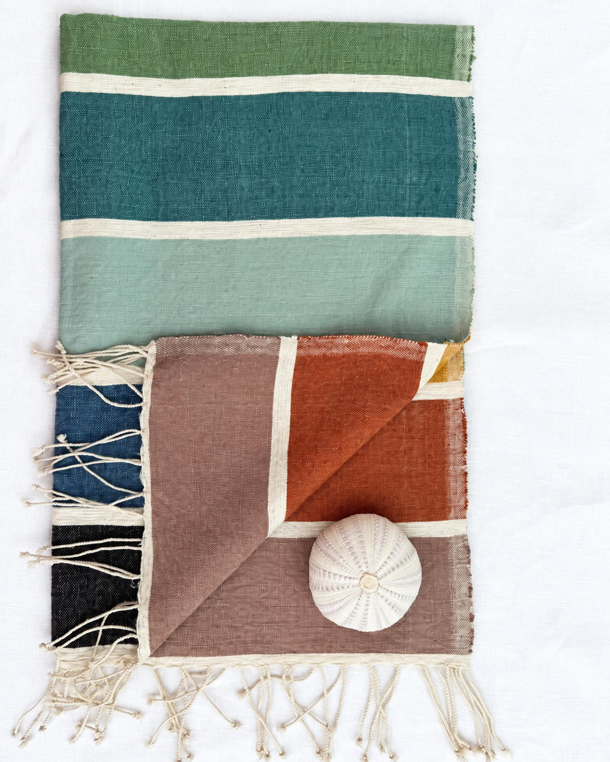 a woven towel laid flat on a white background. The larger textile at the top has horizontal stripes in olive green, teal, mint, and cream.