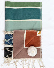 a woven towel laid flat on a white background. The larger textile at the top has horizontal stripes in olive green, teal, mint, and cream.