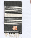 A folded dark gray woven towel with cream and white striped patterns, featuring a small seashell applique and cream fringe tassels at the bottom, laid flat on a white background.