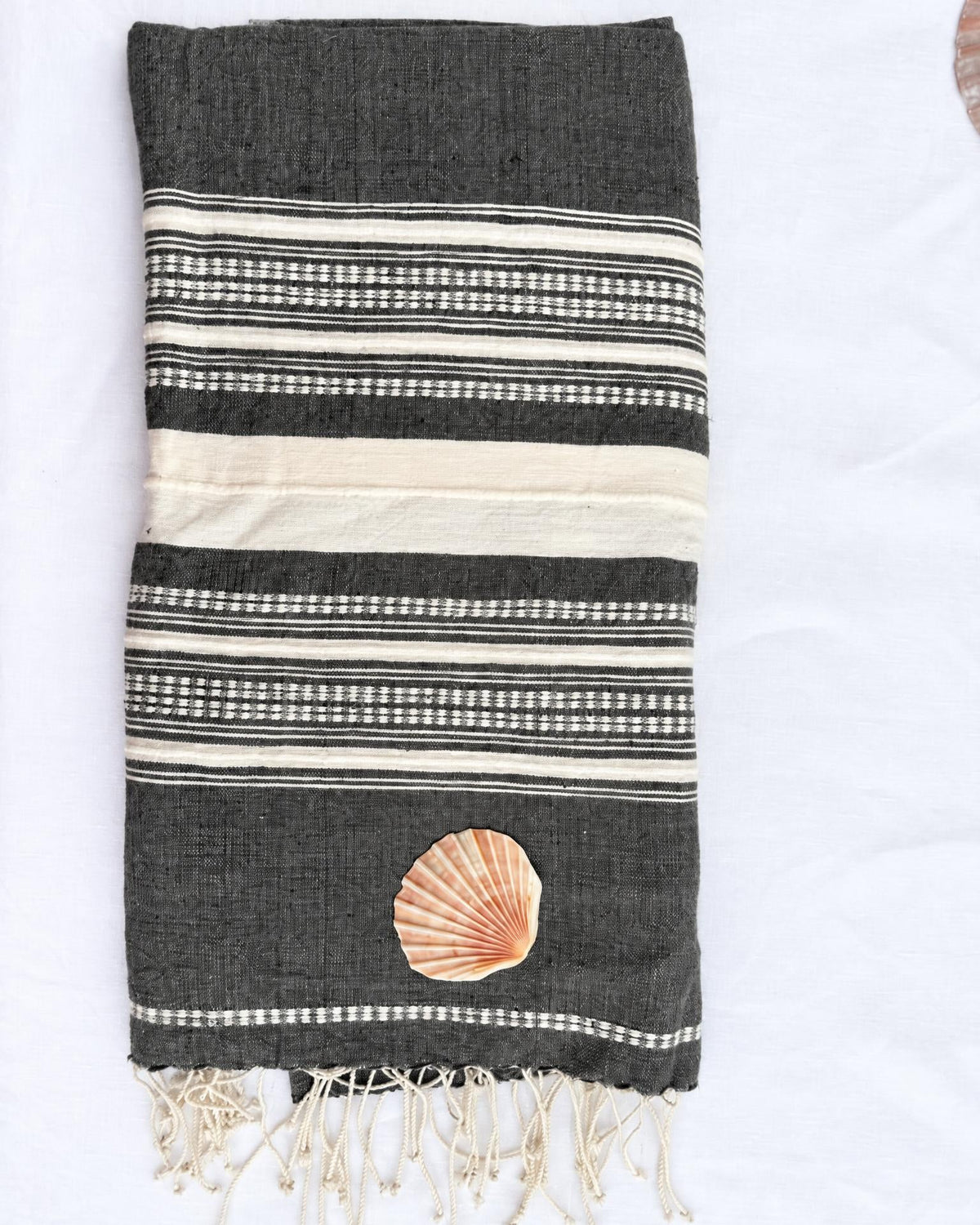 A folded dark gray woven towel with cream and white striped patterns, featuring a small seashell applique and cream fringe tassels at the bottom, laid flat on a white background.