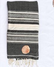 A folded dark gray woven towel with cream and white striped patterns, featuring a small seashell applique and cream fringe tassels at the bottom, laid flat on a white background.
