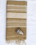 A folded beige and cream striped cotton towel with fringed edges, styled on a white fabric background with a seashell prop.
