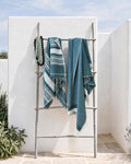 Two teal and white striped woven towels draped over a metal folding rack in an outdoor patio setting with white stucco walls and greenery.