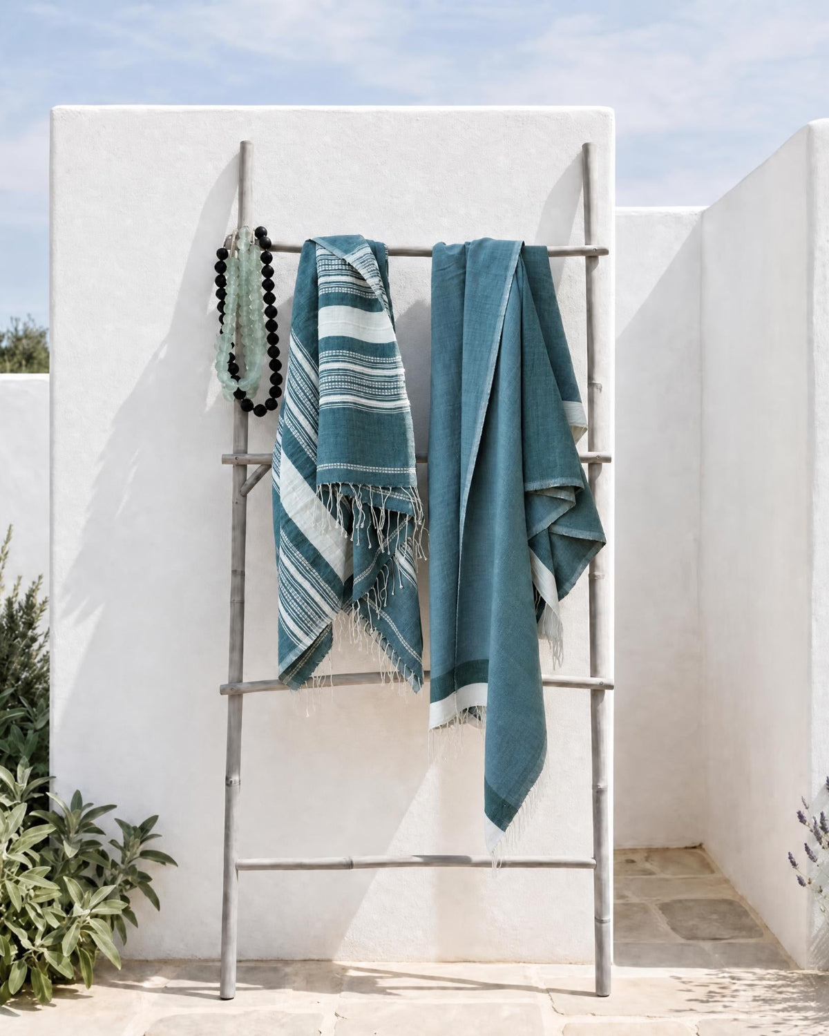 Two teal and white striped woven towels draped over a metal folding rack in an outdoor patio setting with white stucco walls and greenery.