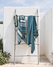 Two teal and white striped woven towels draped over a metal folding rack in an outdoor patio setting with white stucco walls and greenery.