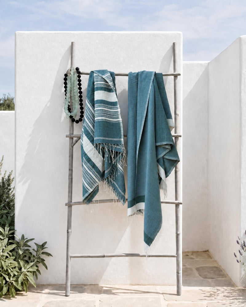 Two teal and white striped woven towels draped over a metal folding rack in an outdoor patio setting with white stucco walls and greenery.