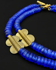 A close-up of a double-strand beaded necklace featuring vibrant blue faceted beads and textured gold-toned spiral accents, displayed on a solid black background.