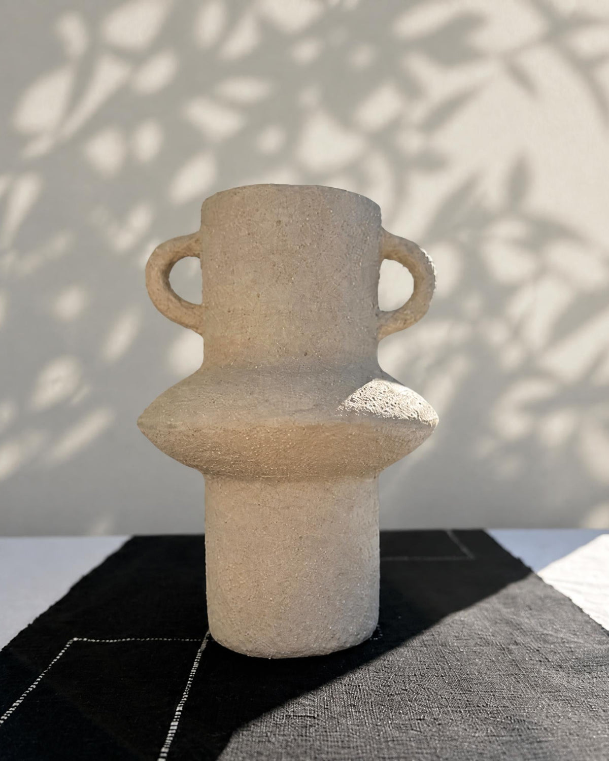 Beige ceramic vase with handles on a textured surface with shadows cast by leaves.
