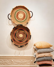 Two round woven baskets with handles are displayed on a cream wall above a decorative molding, with a stack of five textured throw pillows on the right.