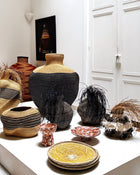 A collection of hand-woven baskets and decorative vases and bowls displayed on a white pedestal in a bright, minimalist room with white paneled doors.