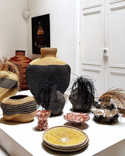 A collection of hand-woven baskets and decorative vases and bowls displayed on a white pedestal in a bright, minimalist room with white paneled doors.