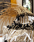A close-up of a handwoven basket with a natural tan base, black and white woven stripes, and white fringed tassels, with dried grasses on top.