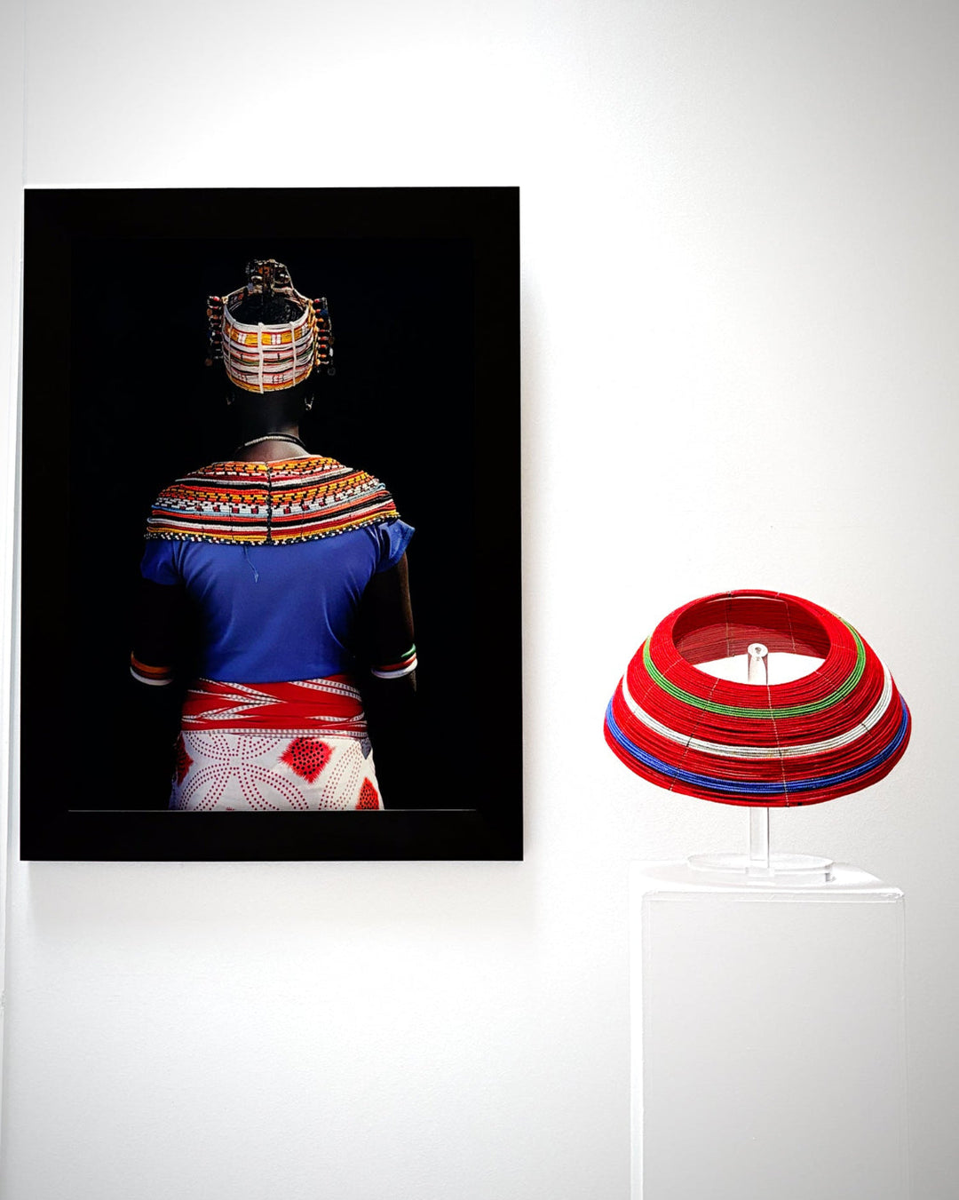 A framed art print depicting a person from behind wearing a vibrant blue, red, and white patterned traditional outfit with a beaded headpiece, displayed on a white wall next to a red, white, green, and blue striped conical hat on a clear acrylic stand.