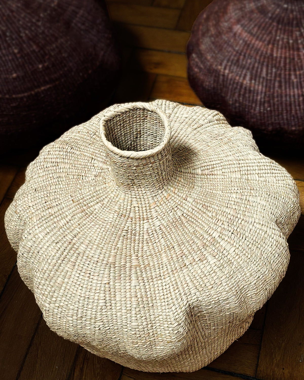 A hand-woven natural fiber vase with a flared, rounded body and a short neck, resting on a wooden floor with other woven baskets in the background.
