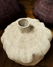 A hand-woven natural fiber vase with a flared, rounded body and a short neck, resting on a wooden floor with other woven baskets in the background.